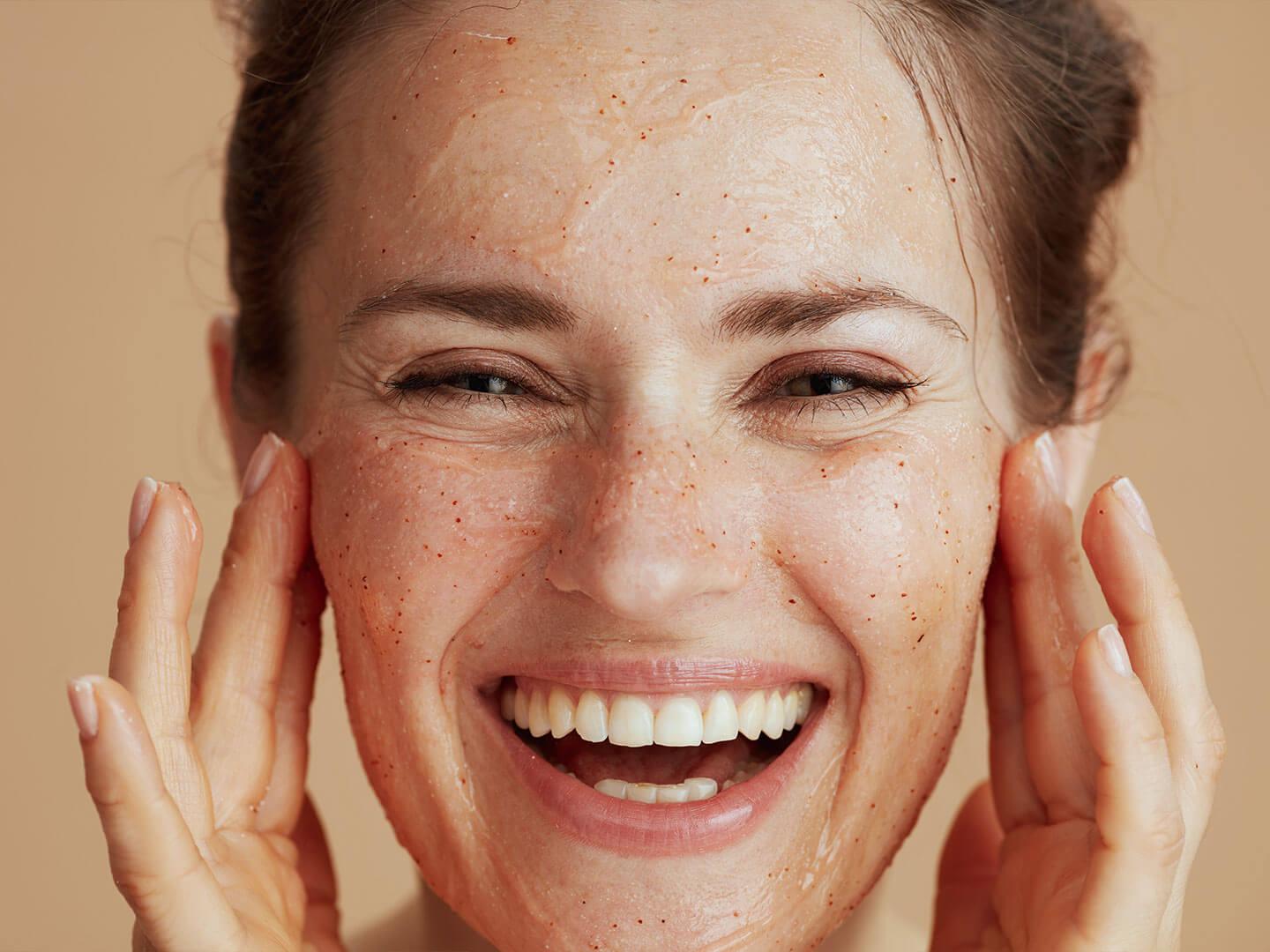 An image of a happy woman, smiling widely infront of the camera while she gently scrubs a face exfoliator onto her skin Desktop