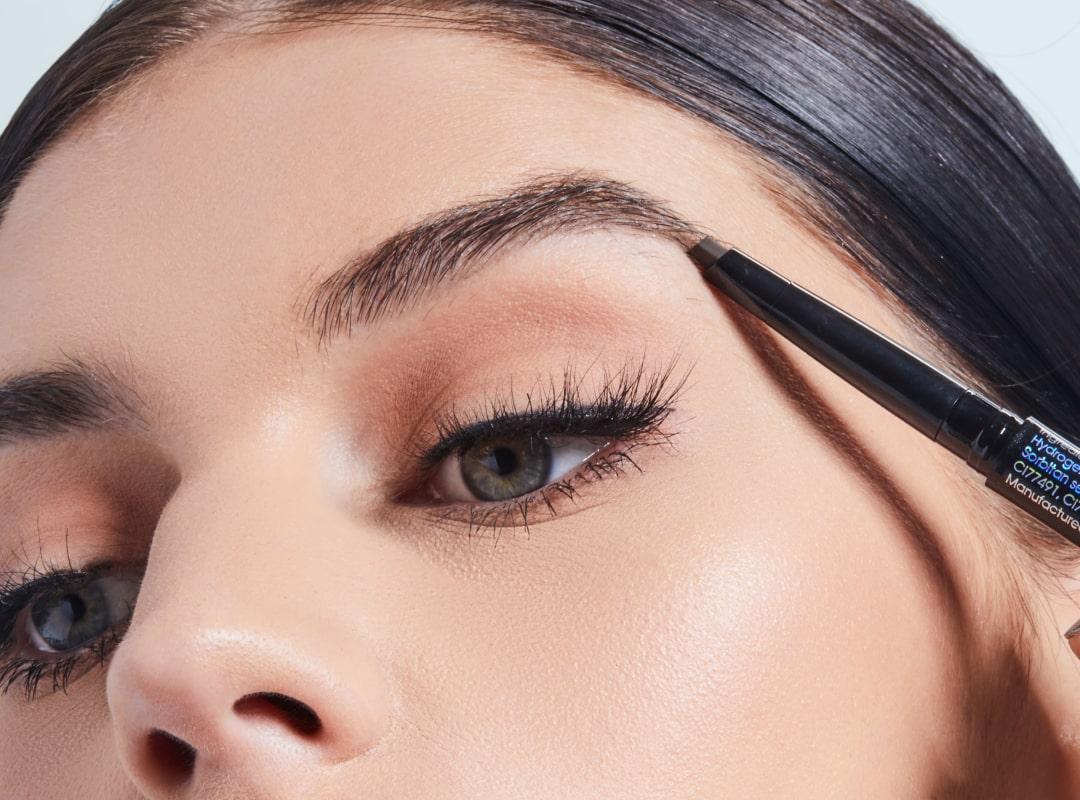 A close-up photo of a woman using a browpencil to fill in her eyebrows while wearing black eyeliner