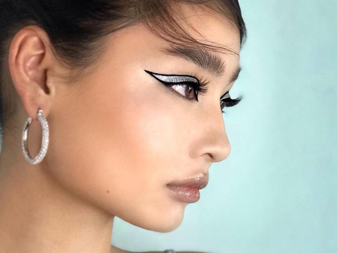 An image of a young woman striking a side-profile pose, elegantly adorned with black eyeliner and shimmering silver eyeshadow wearing silver hoop earrings and with a glossy lip finish