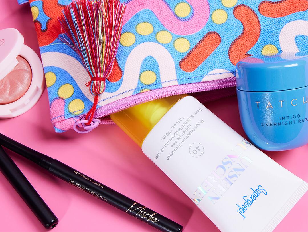 Beauty products from various brands and colorful makeup pouch with tassel on pink background