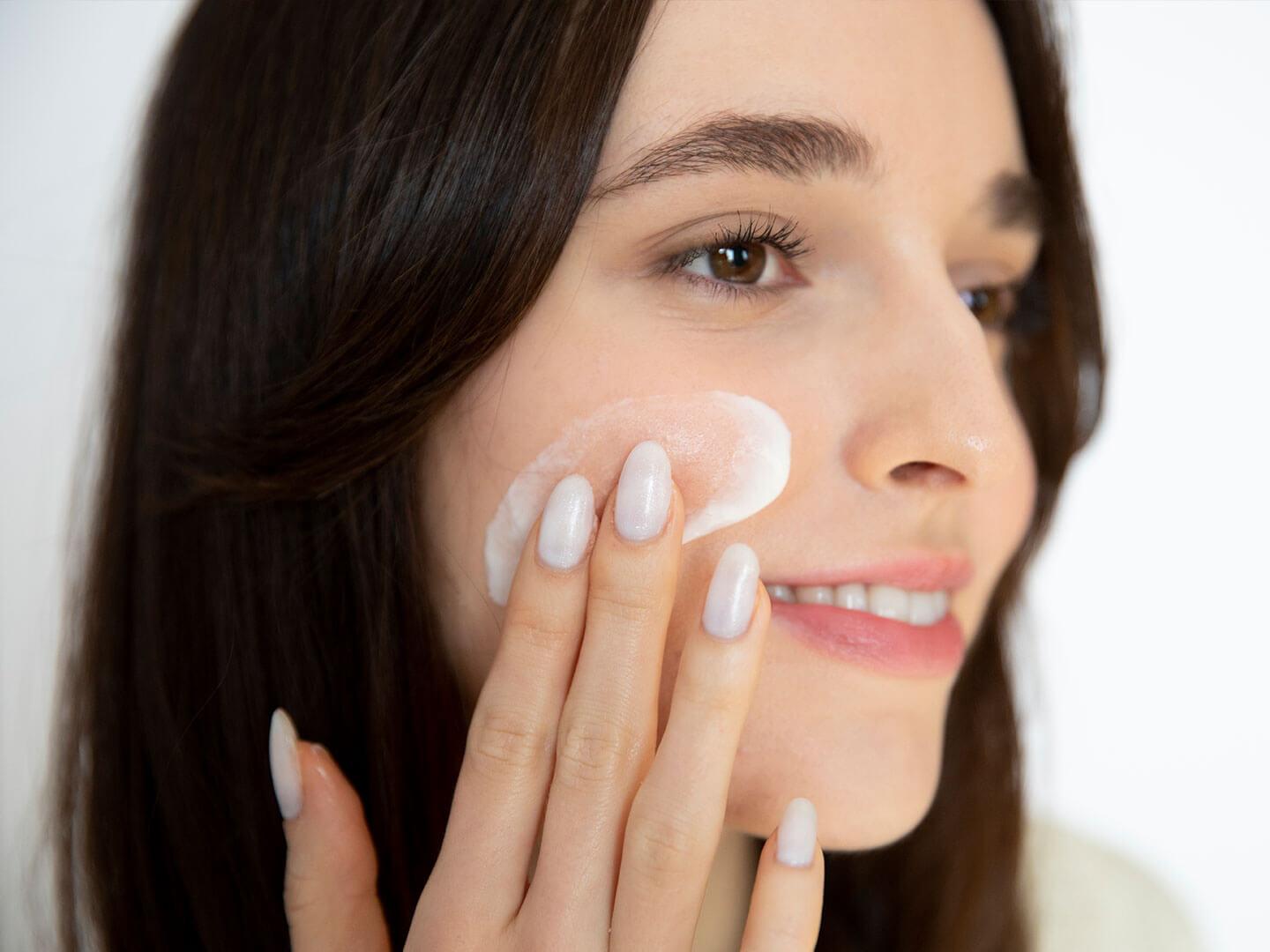 An image of a woman with clear skin, her nails manicured in a white nail polish applying a moisturizer while smiling sideways Desktop