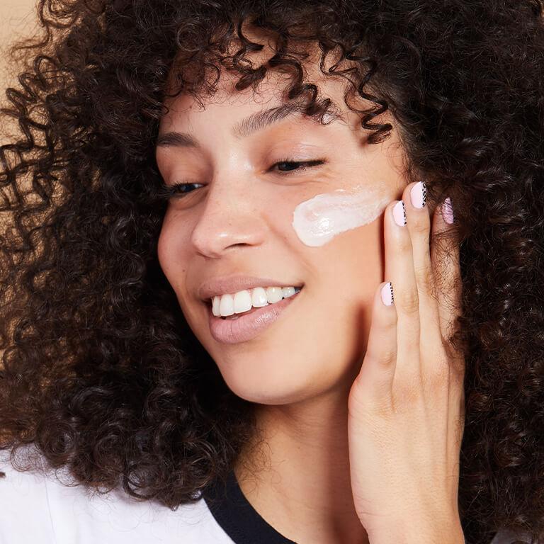 An image of a smiling model with curly hair applying cream on her cheek Desktop