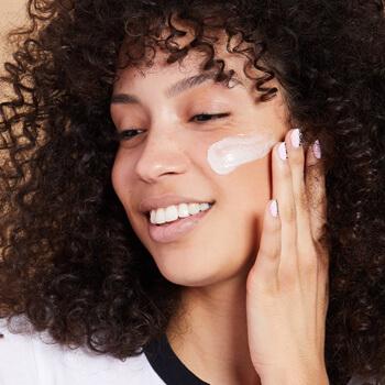 An image of a smiling model with curly hair applying cream on her cheek Mobile